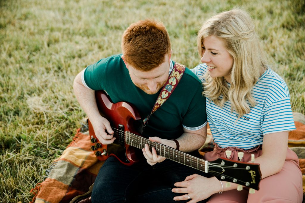 Couple sit together while groom plays guitar at golden hour pre wedding shoot warwickshire